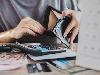Pessoa abrindo carteira vazia em cima de uma mesa cheia de notas de dinheiro, calculadora, cartões de crédito e contas