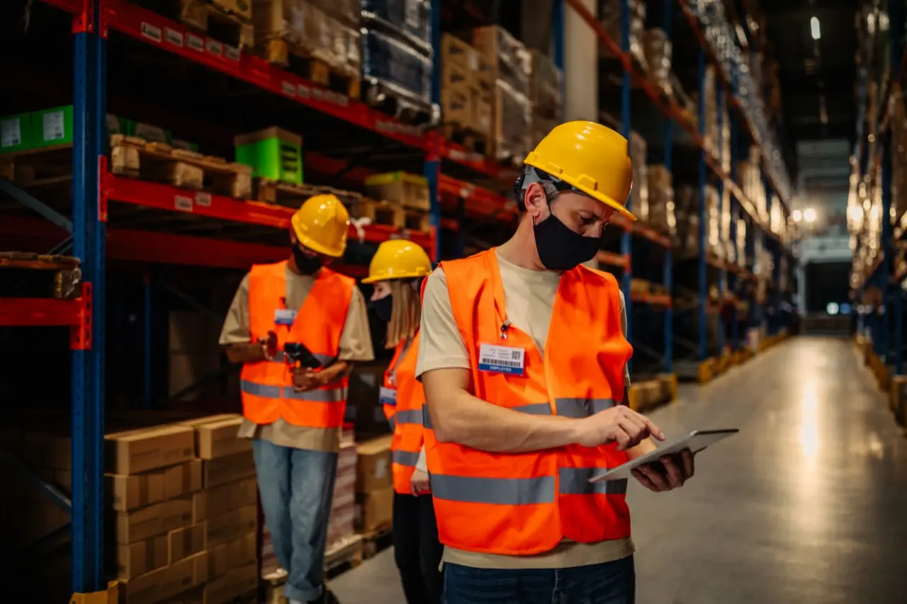 Três pessoas de capacete de segurança, colete refletivo e máscara facial conferem caixas em um estoque