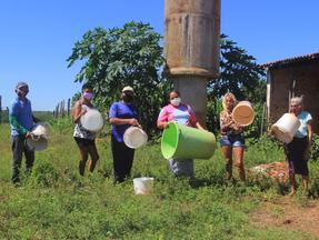 Em frente a uma caixa d'água/chafariz, famílias mostram baldes vazios, denunciando escassez. Projeto de abastecimento hídrico não foi concluído.
