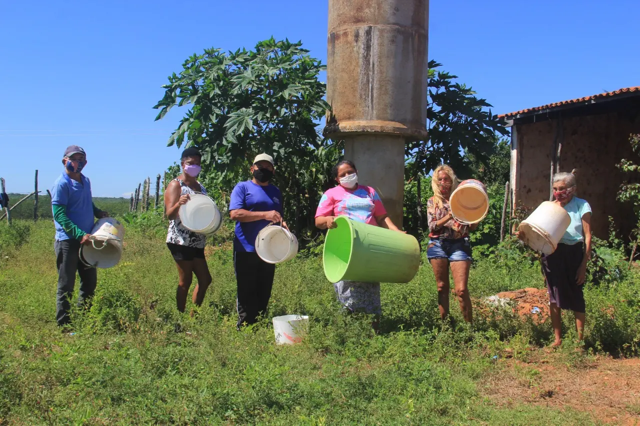Em frente a uma caixa d'água/chafariz, famílias mostram baldes vazios, denunciando escassez. Projeto de abastecimento hídrico não foi concluído.