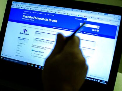 Sombra de mão segurando caneta em frente a notebook mostrando o site da Receita Federal