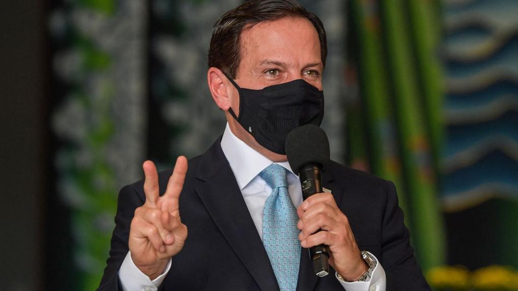 Governador de São Paulo João Doria fazendo símbolo de 2 com o dedo e segurando um microfone durante evento
