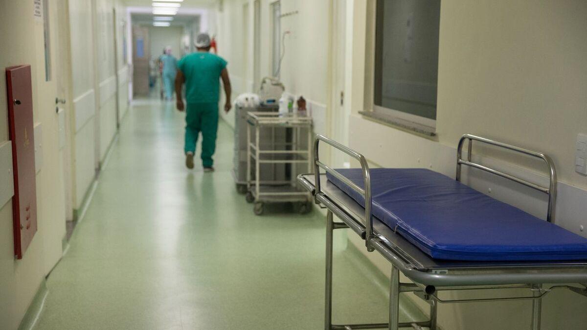 Corredor de um hospital, com uma maca vazia, e um profisisonal caminha ao fundo