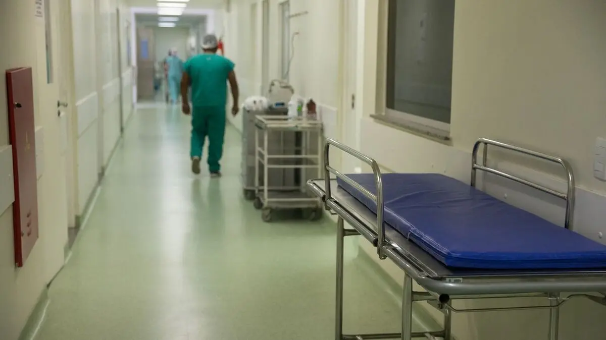 Corredor de um hospital, com uma maca vazia, e um profisisonal caminha ao fundo