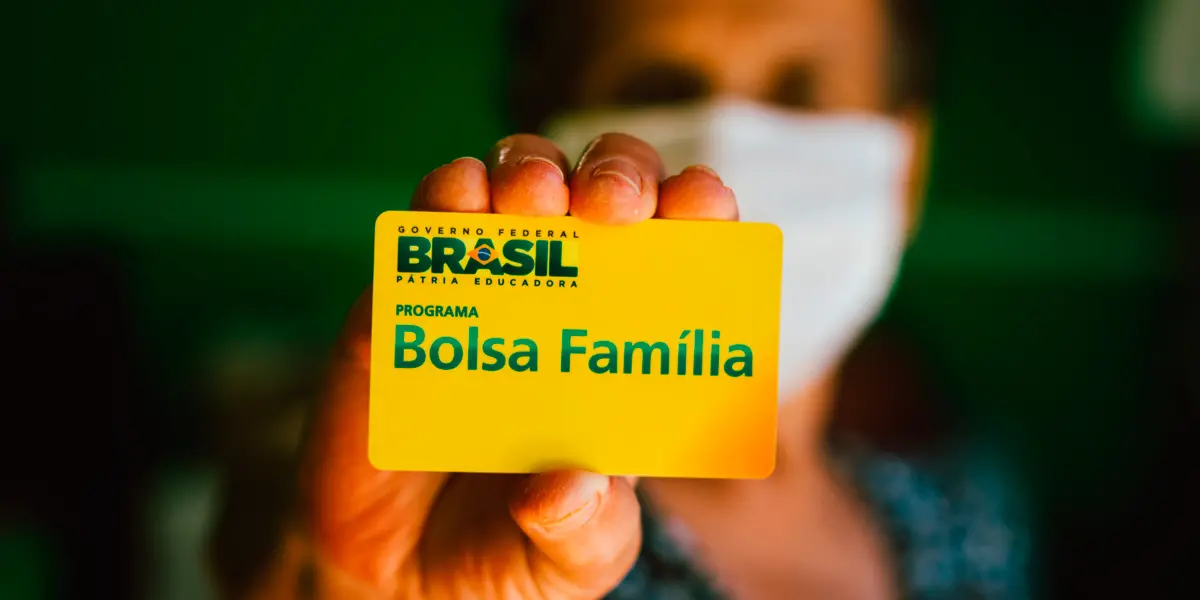 Mulher de máscara segura um cartão do Bolsa Família