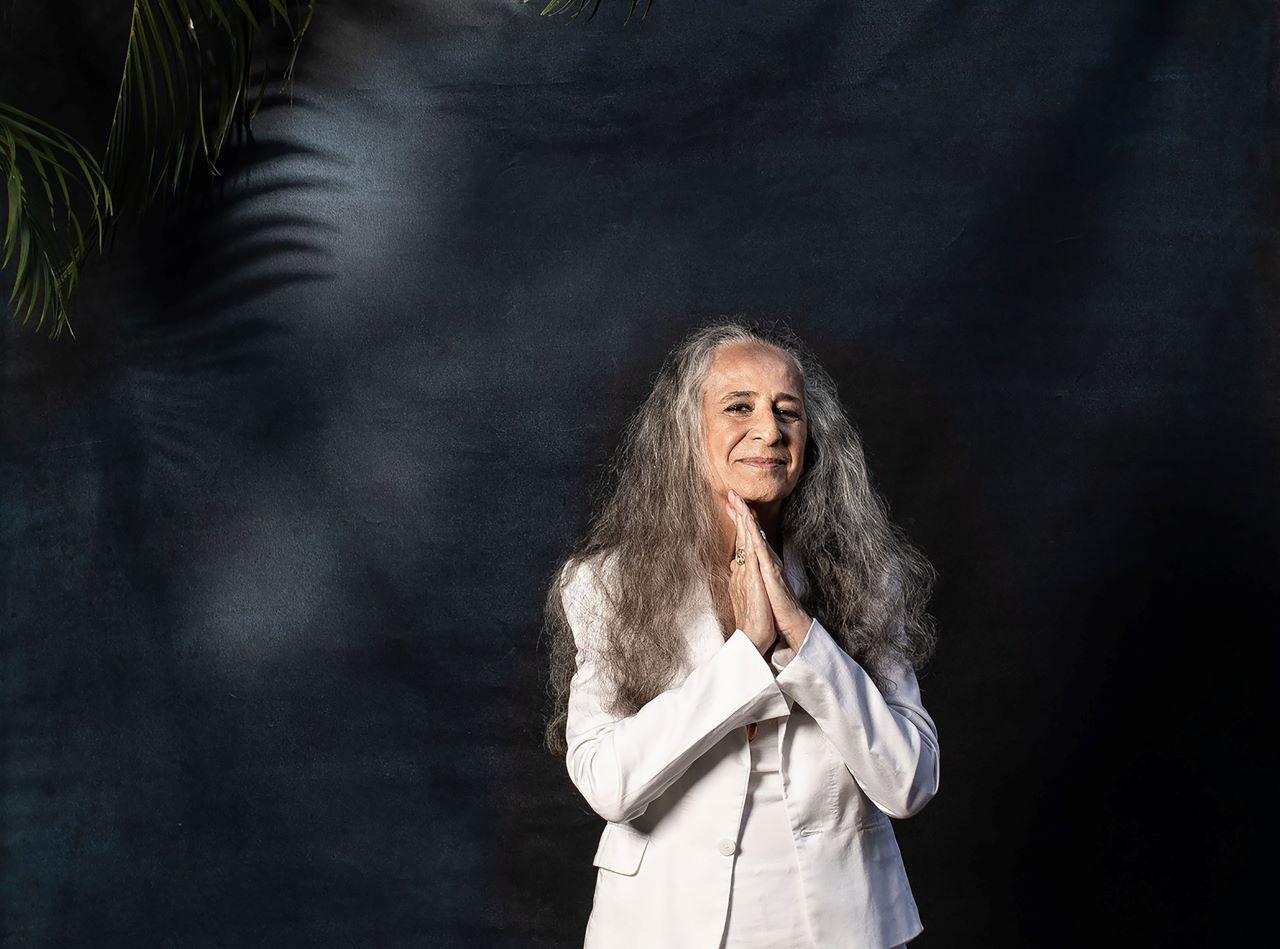 maria bethânia de branco, com as mãos postas, diante de uma parede escura