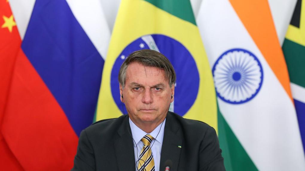 Bolsonaro com semblante sério. Bandeira do Brasil ao fundo