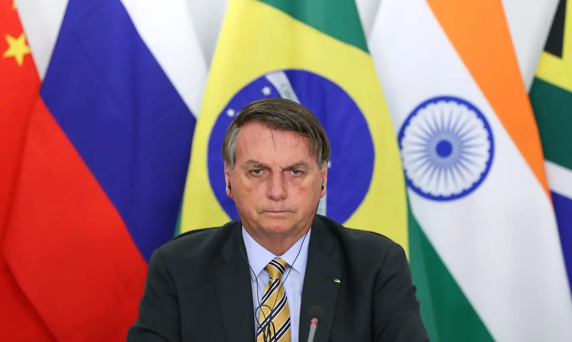 Bolsonaro com semblante sério. Bandeira do Brasil ao fundo