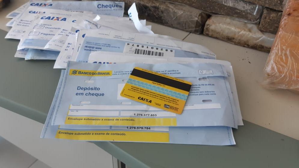 Cartão do auxílio emergencial sobre envelopes da Caixa e com drogas ao redor