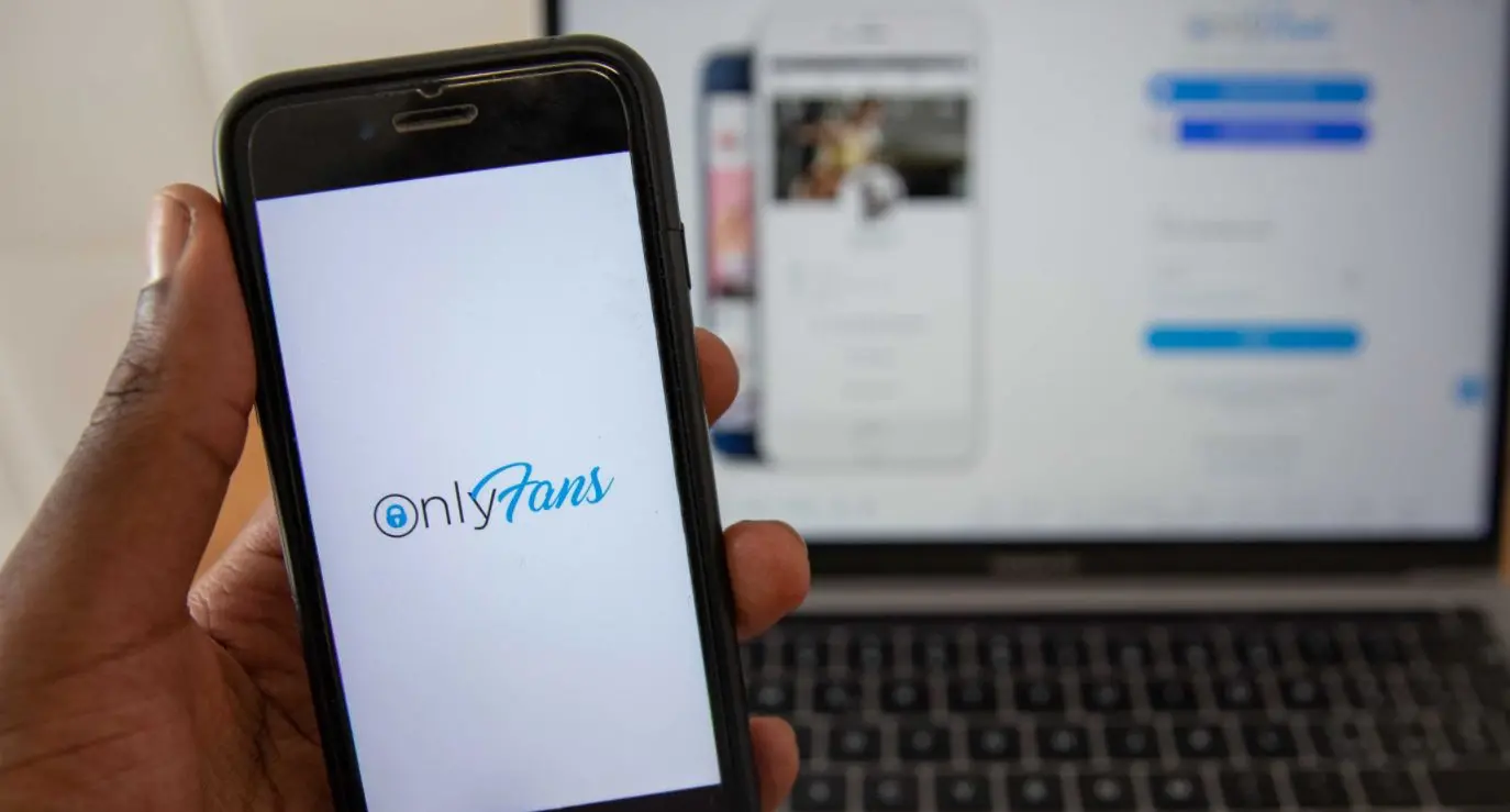 Celular com logo do OnlyFans sendo segurado em frente a notebook