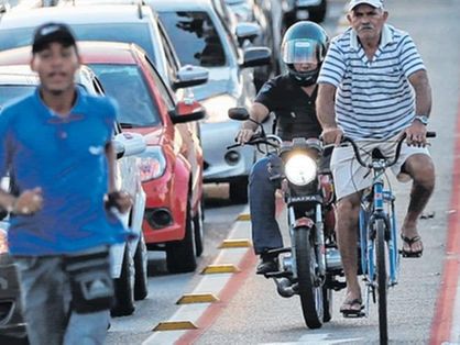 Foto do trânsito em Fortaleza, no Ceará, mostra vendedor ambulante de boné e blusa azul correndo perto dos veículos parados em uma via. Na imagem, motociclista é visto na ciclofaixa, onde um ciclista está pedalando