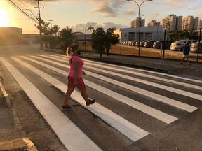Mulher loira de cabelo amarrado usando máscara de proteção facial, blusa e short rosa e tênis preto atravessando faixa de pedestres em Fortaleza, no Ceará