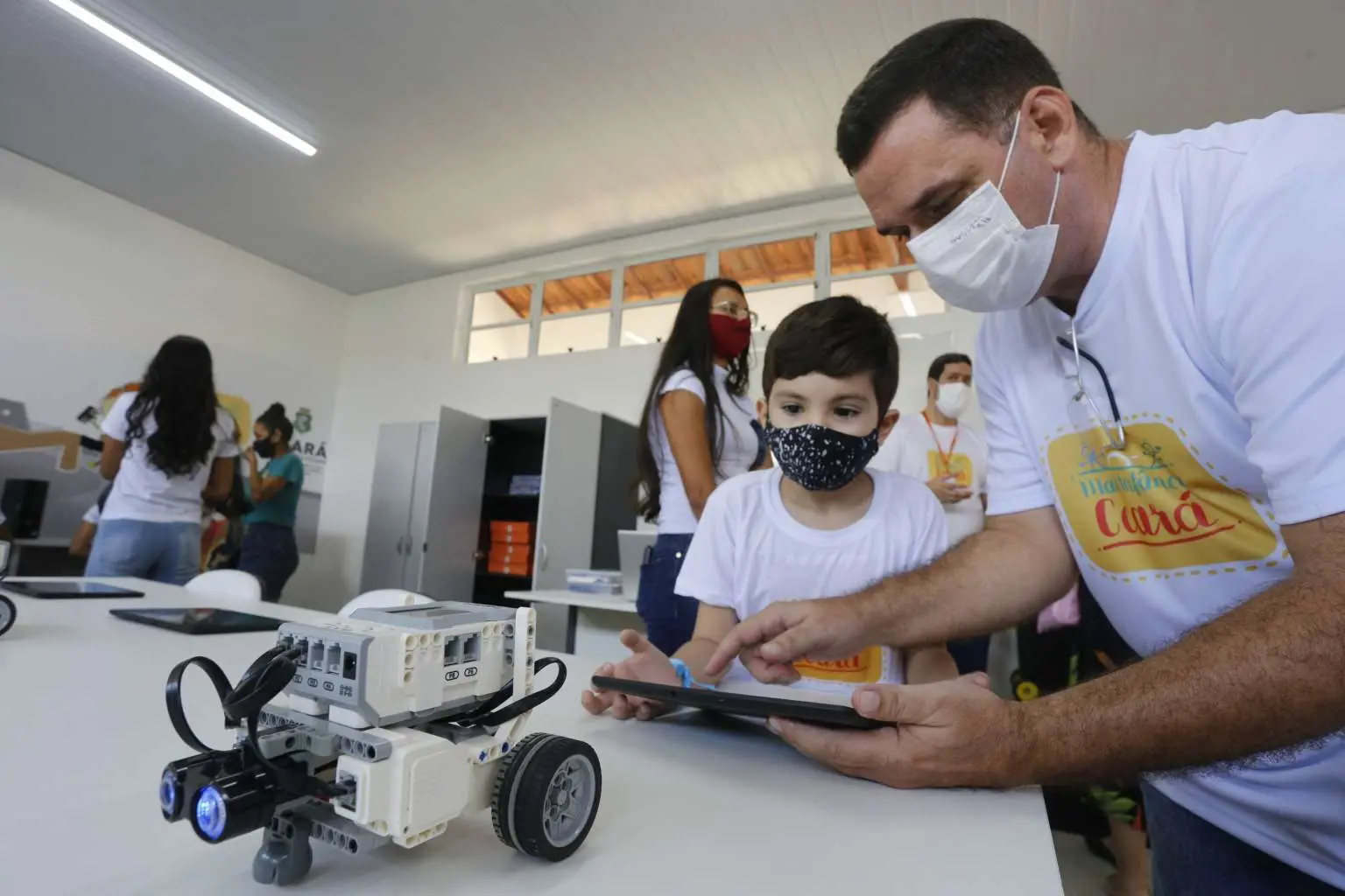 Imagem mostra uma criança de máscara e um adulto também de máscara numa sala onde será ministrado o curso de robótica.