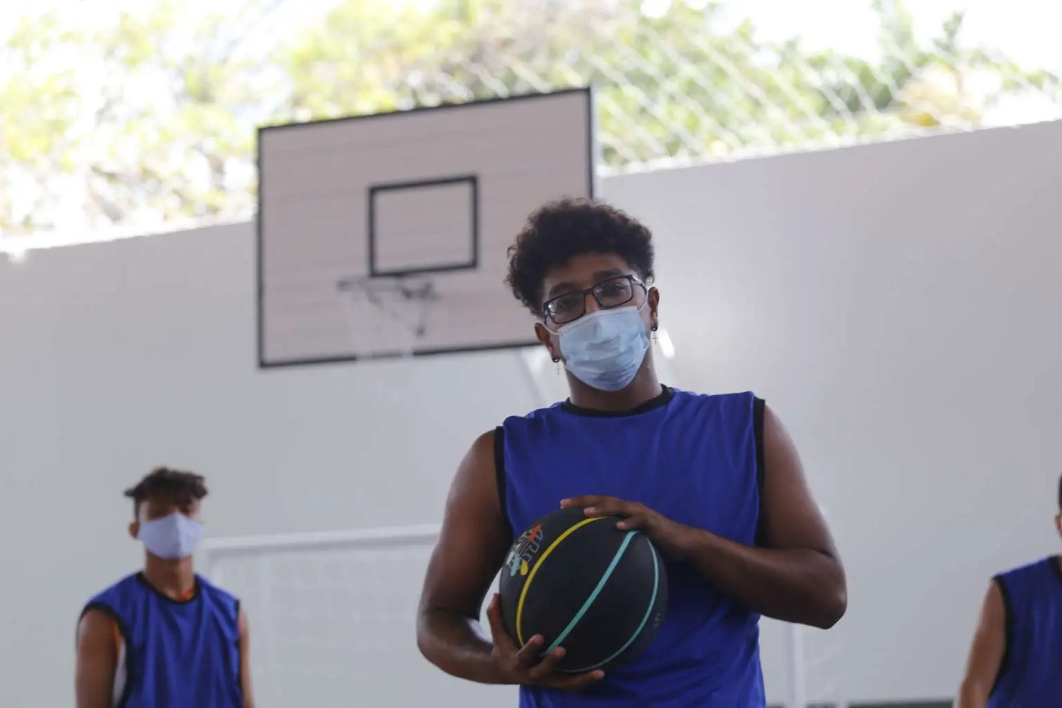 Imagem mostra dois adolescentes de máscara em uma quadra de basquete. Um deles, negro, de óculos, está em evidência, segurando uma bola.