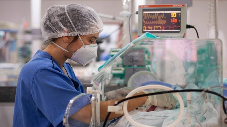 Profissional da saúde com máscara, touca e bata azul em uma UTI neonatal