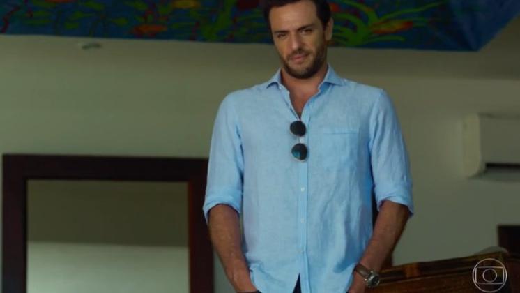 A imagem mostra Rodrigo Lombardi como o empresário Alex, de Verdades Secretas. Ele está num resort, vestido com uma camisa social azul.