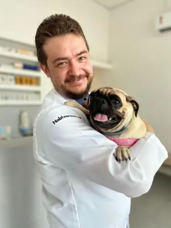 Médico veterinário Dr.Bovy aparece na foto de bata, sorrindo e segurando um pug