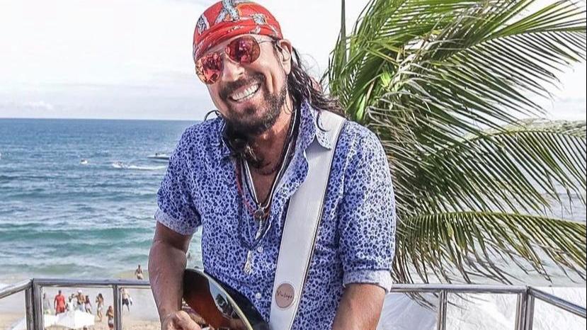 O cantor baiano de axé Bell Marques está numa praia, de bandana vermelha e camisa azul, segurando uma guitarra.