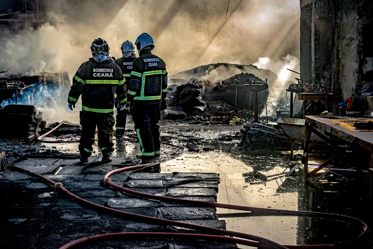 Bombeiros paramentados em meio à fumaça e chamas, após controlarem o fogo