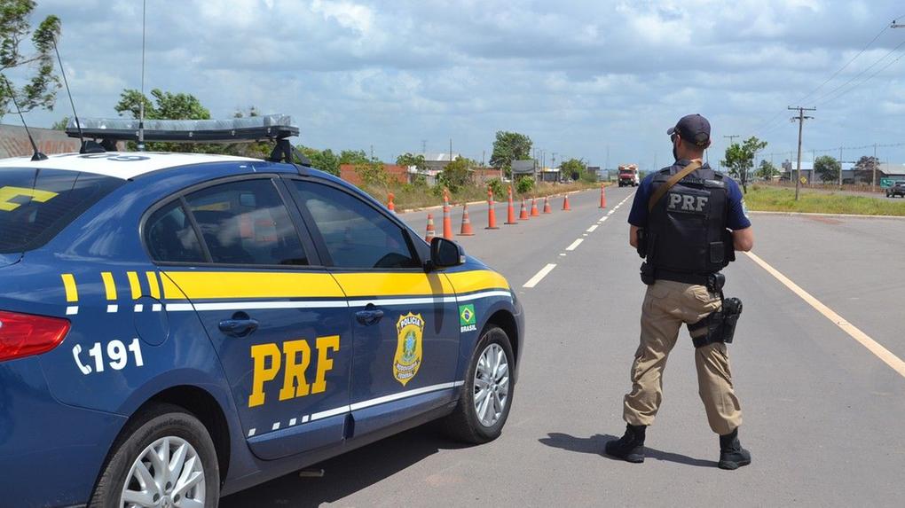 A imagem mostra um agente da Polícia Rodoviária Federal em meio a uma rodovia ao lado de uma viatura.