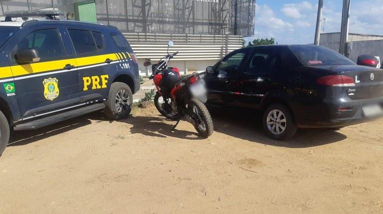 A imagem mostra uma viatura da Polícia Rodoviária Federal ao lado de uma moto e de um carro que foram apreendidos.