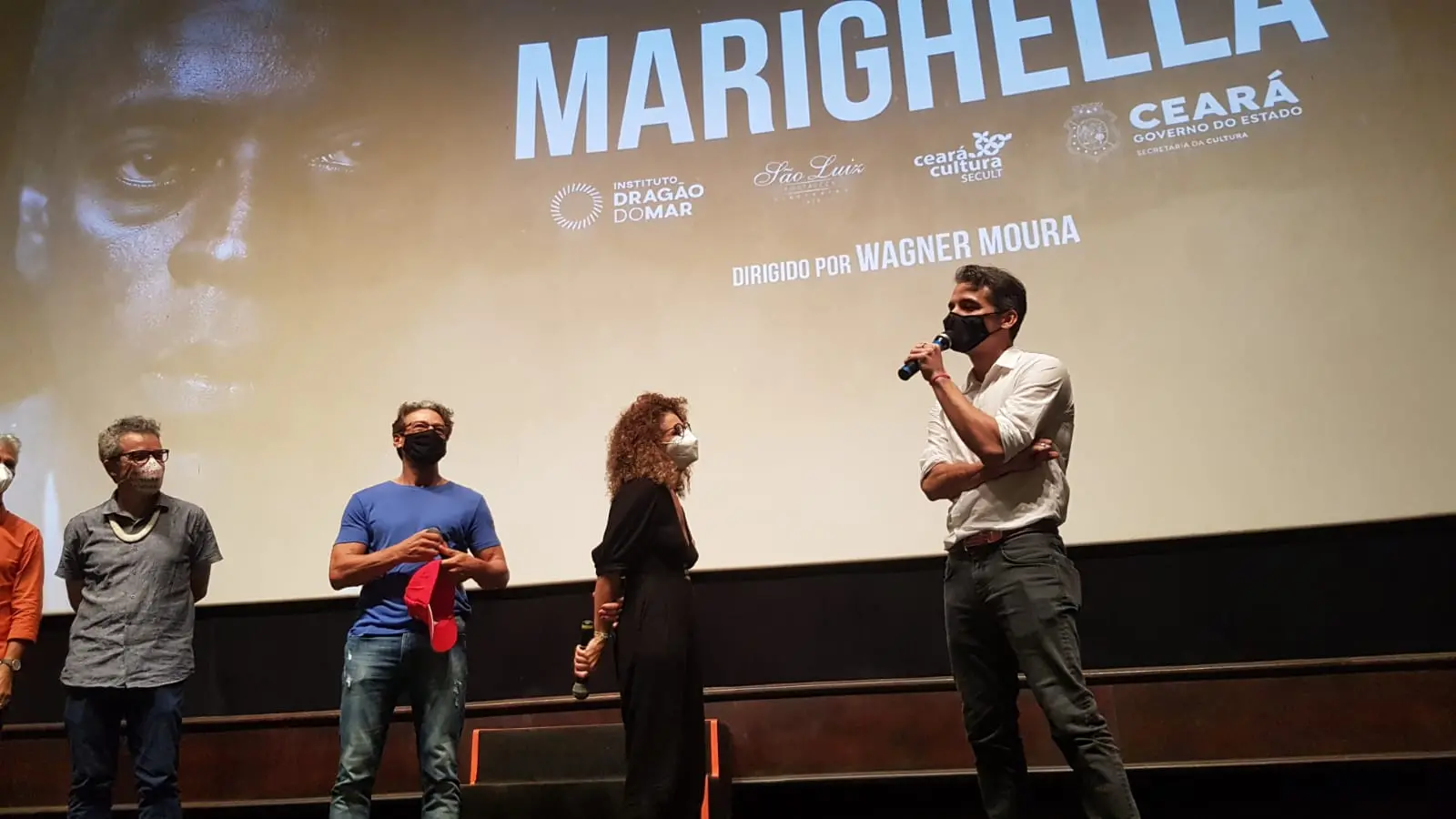 wagner moura ao microfone ao lado de outras pessoas. atrás dele, a tela do cinema destaca o nome marighella