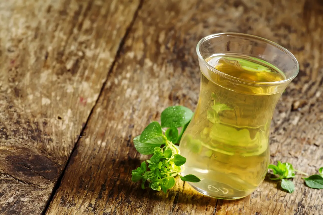 Copo com chá de orégano e folhas de orégano dispostas ao lado sobre uma mesa