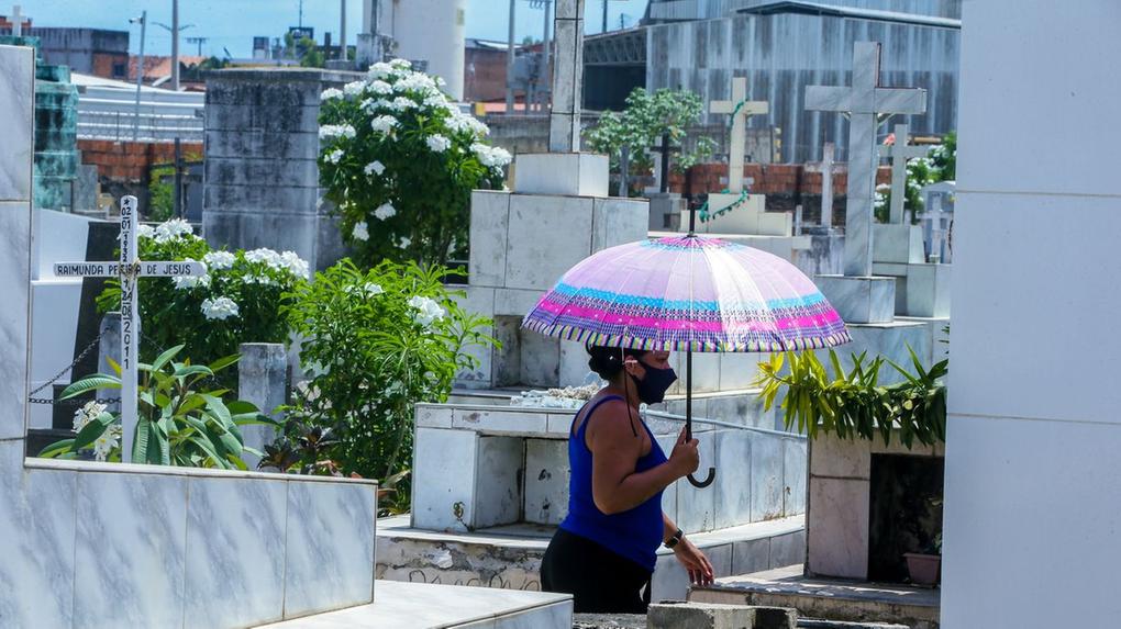 Mulher de máscara e guarda-chuva passeia por entre lápides num cemitério de Fortaleza.