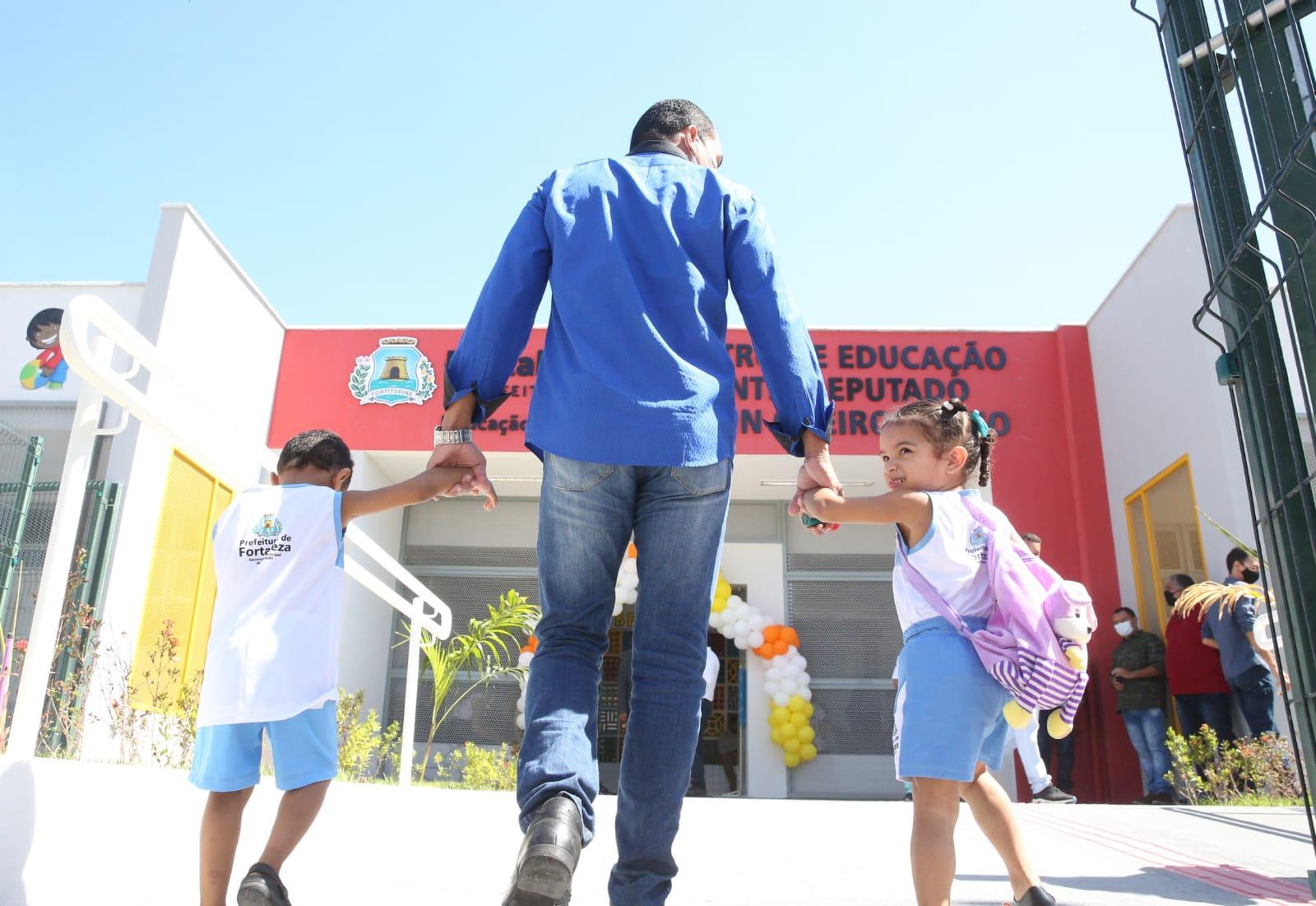 De mãos dadas, duas crianças entram na escola acompanhadas de um responsável
