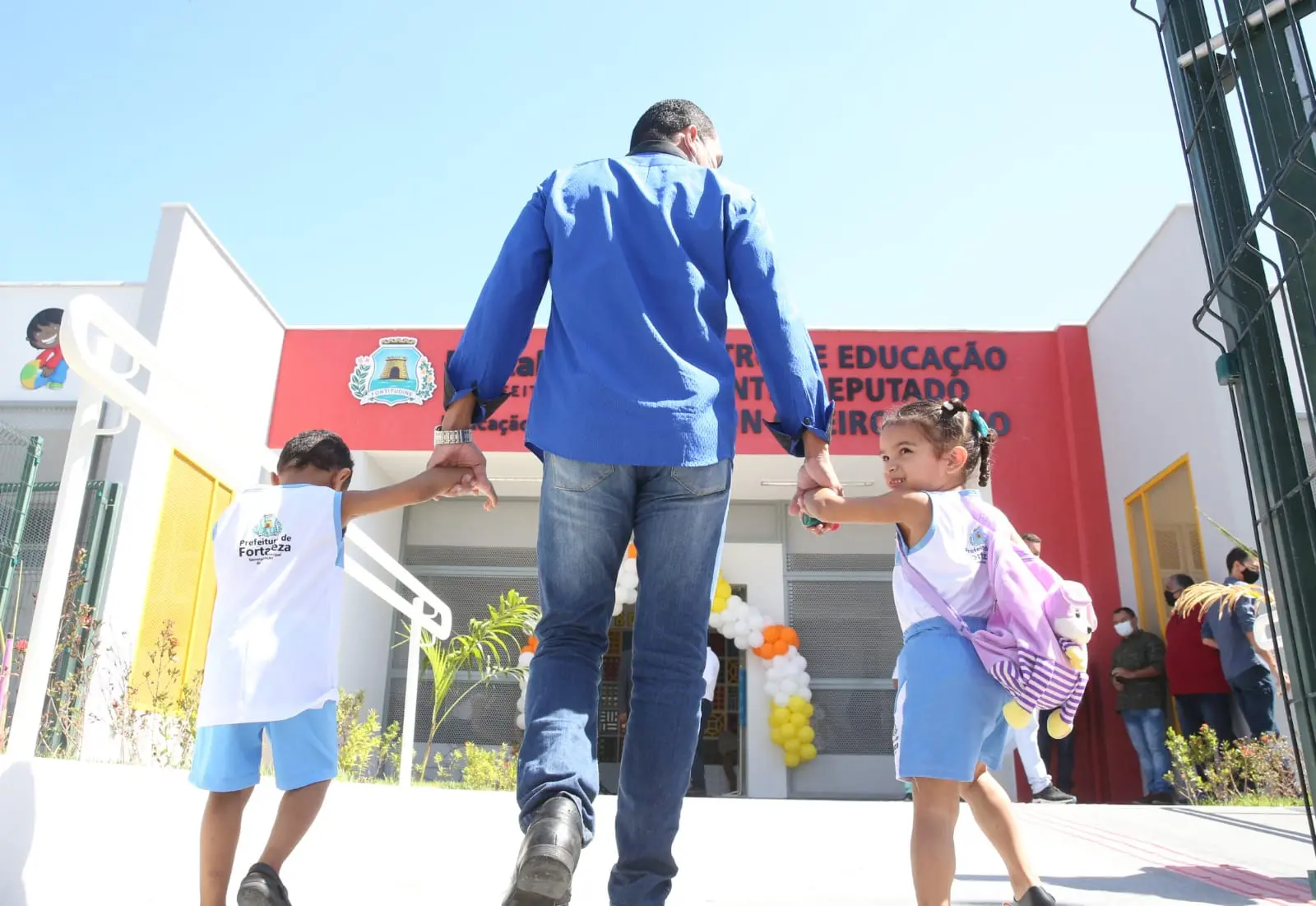 De mãos dadas, duas crianças entram na escola acompanhadas de um responsável