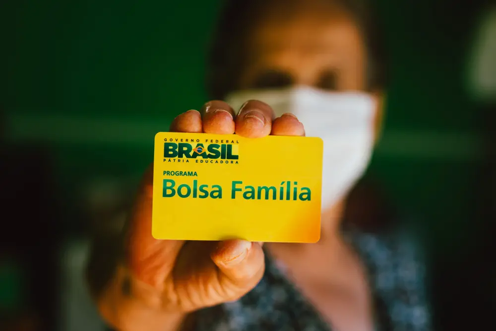 Mulher ao fundo mostra cartão do Bolsa Família.