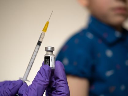 Mão com luva segura dose de vacina e injeção, enquanto criança aparece atrás, embaçada