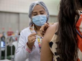 Profissional da saúde aplica injeção no braço de um mulher, que aparece de costas