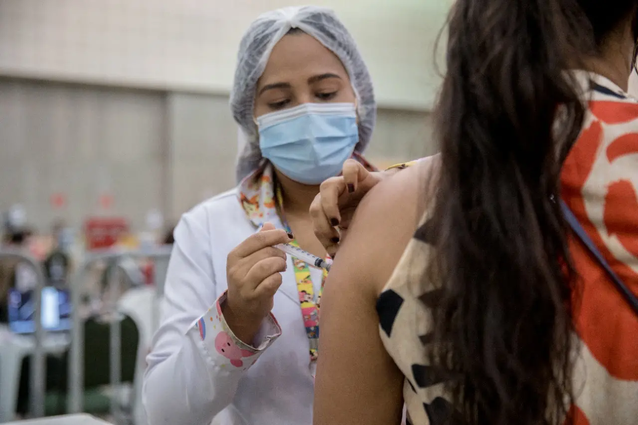 Profissional da saúde aplica injeção no braço de um mulher, que aparece de costas