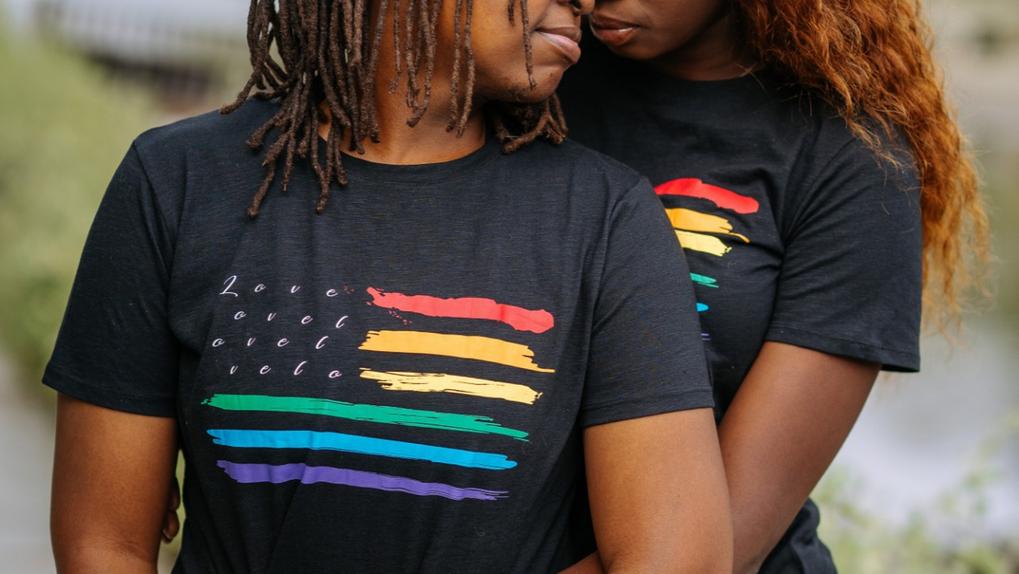 Casal LGBTI+ abraçado, vestindo camisa com bandeira do movimento