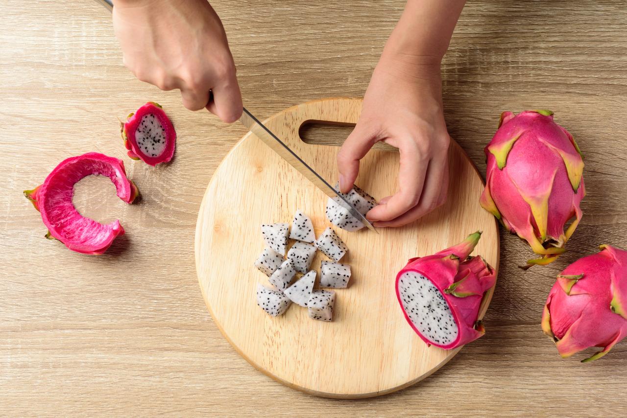 Mãos fatiam uma pitaya sobre uma tábua de madeira ao lado de pitayas interias
