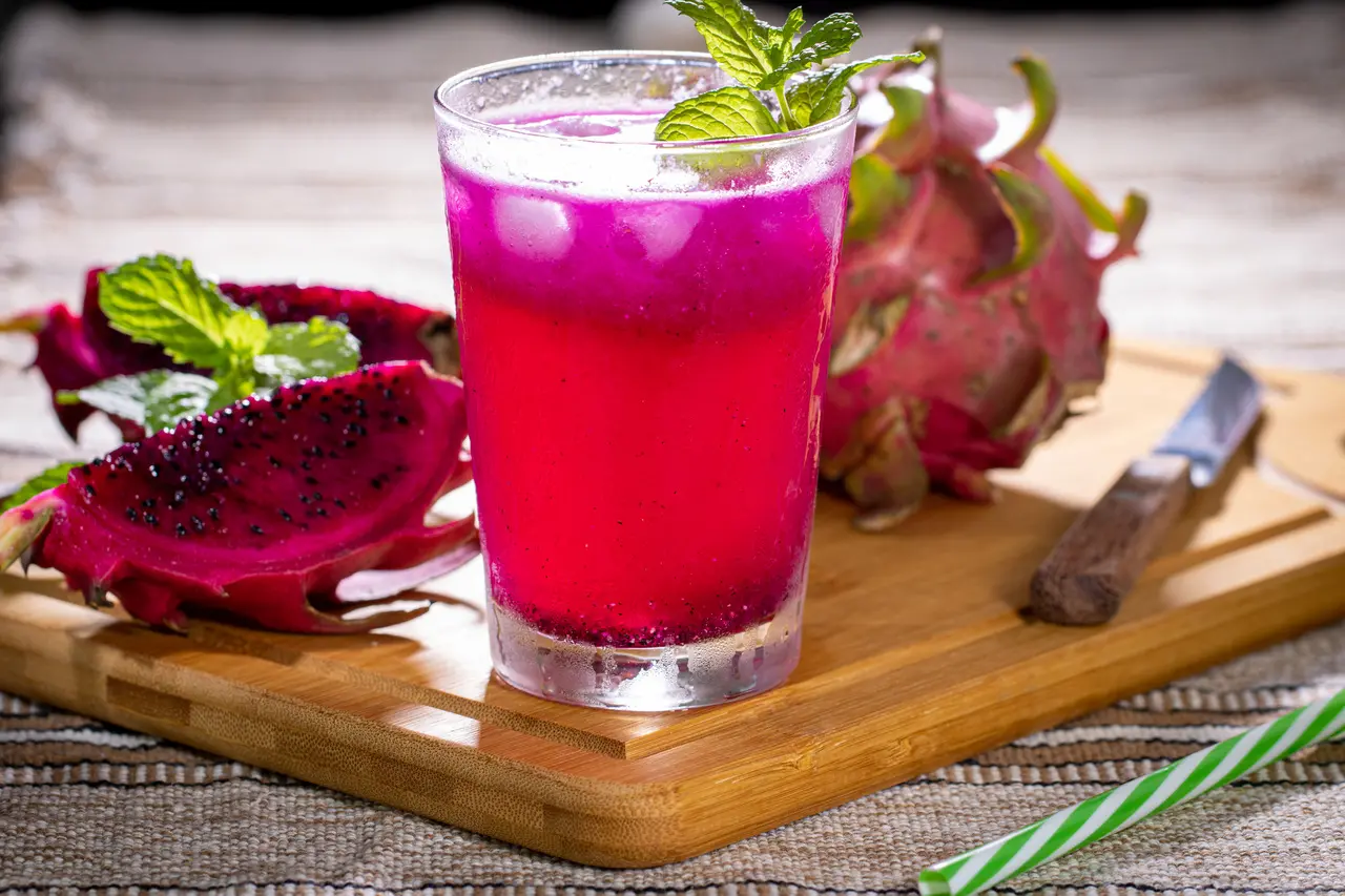 Copo com suco de pitaya e gelo sobre uma tábua de madeira ao lado de fatias de pitaya e um faca