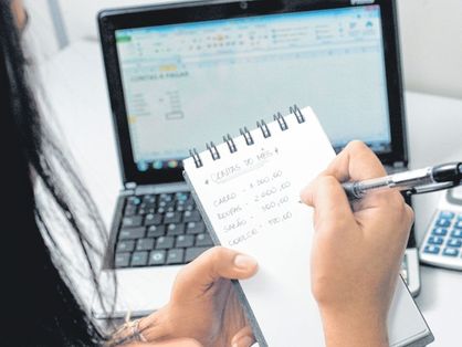 Imagem mostra uma mulher de frente para um notebook e uma calculadora anotando suas dívidas em um bloco notas.