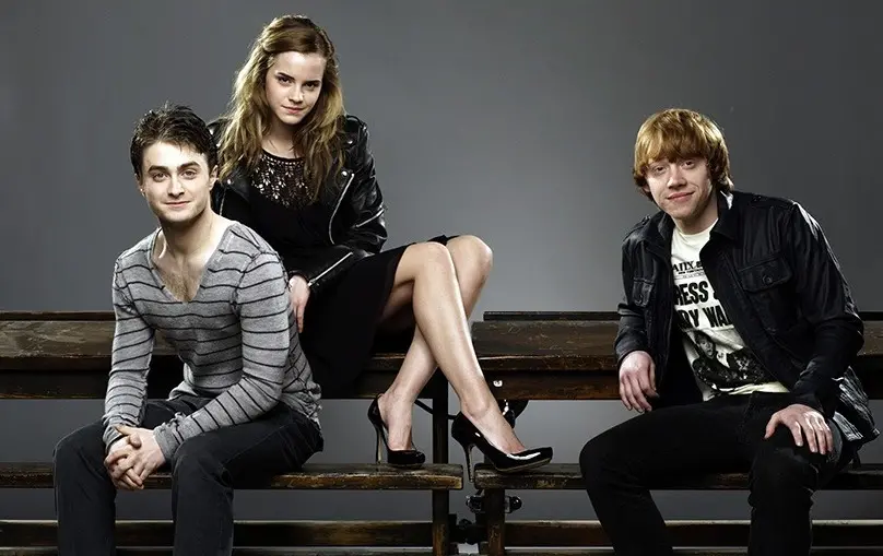 Os atores Daniel Radcliffe, Emma Watson e Rupert Grint numa sessão fotográfica. Eles estão sentados numa cadeira de madeira.