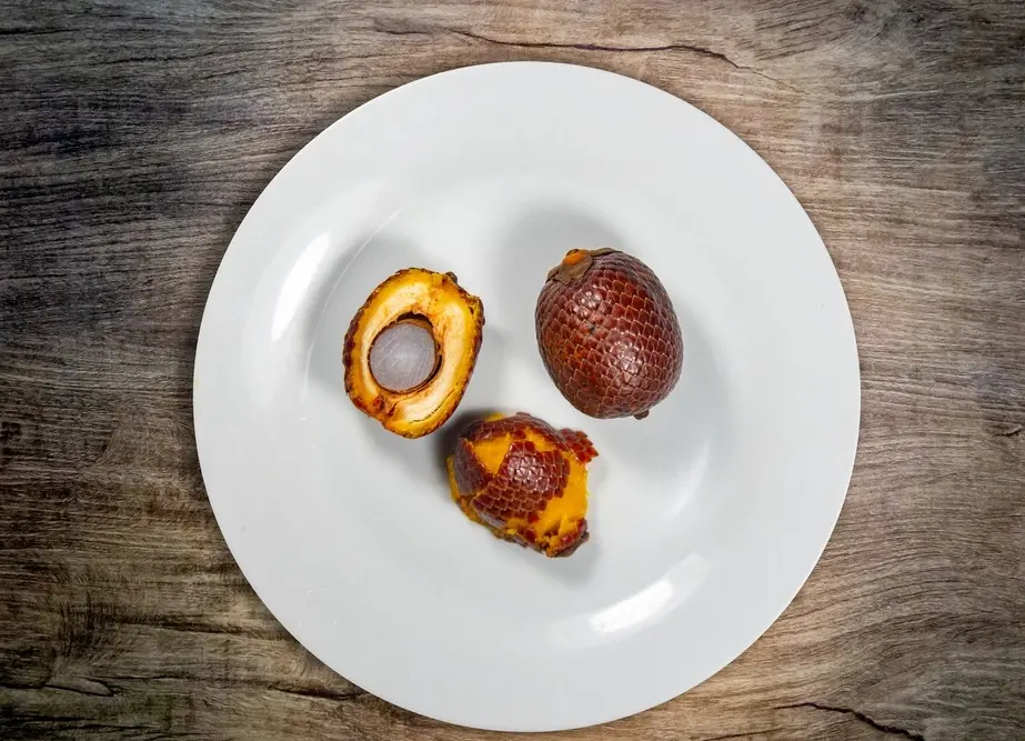 Três buritis, um totalmente com casca, outro sendo descascado e outro cortado, mostrando a amêndoa.