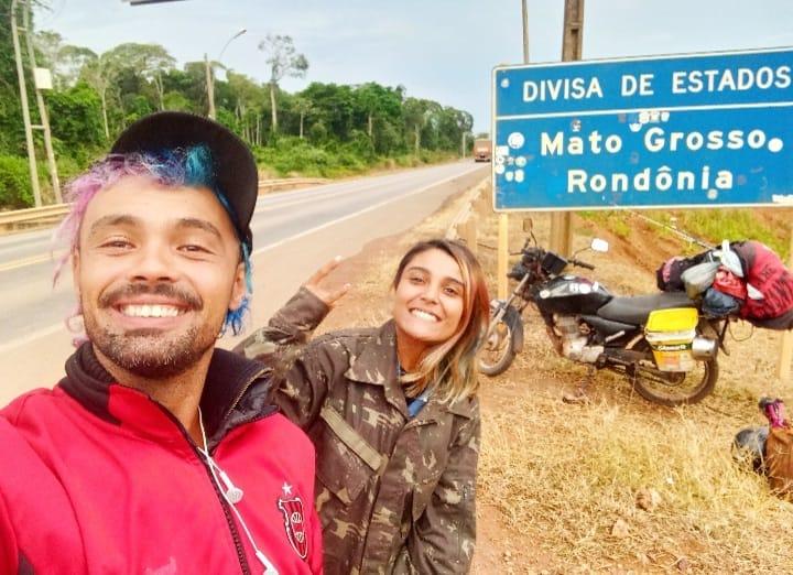 Tiago Bohrer e Jennifer Santos em frente à moto do casal e a uma placa da divisa dos estados de Mato Grosso e Rondônia