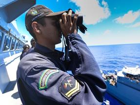 Oficial da Marinha, usando uniforme e boné, observa o mar com um binóculo de um navio da corporação