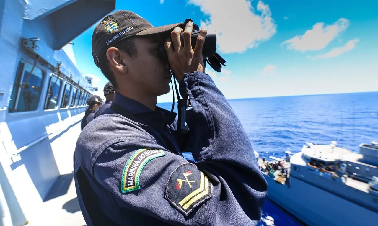 Oficial da Marinha, usando uniforme e boné, observa o mar com um binóculo de um navio da corporação