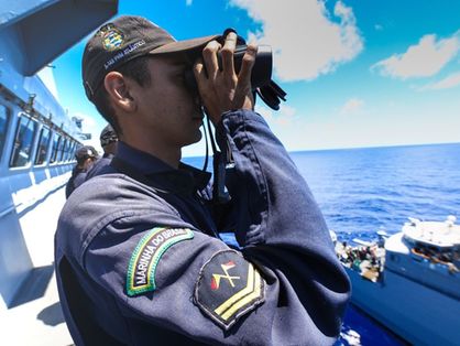 Oficial da Marinha, usando uniforme e boné, observa o mar com um binóculo de um navio da corporação