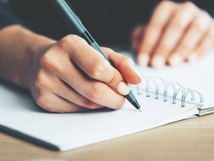 A imagem mostra uma mão escrevendo com uma caneta em um caderno.
