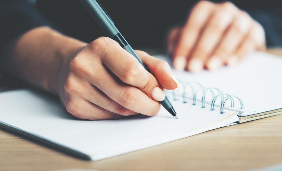 A imagem mostra uma mão escrevendo com uma caneta em um caderno.