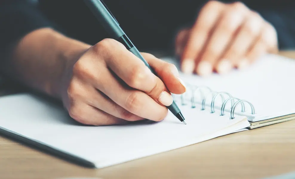 A imagem mostra uma mão escrevendo com uma caneta em um caderno.