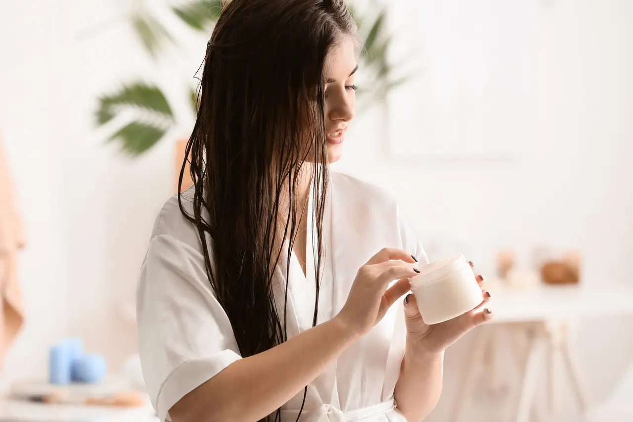 Mulher com cabelo molhado, usando um roupão, pega creme de tratamento capilar com a mão em um cenário branco