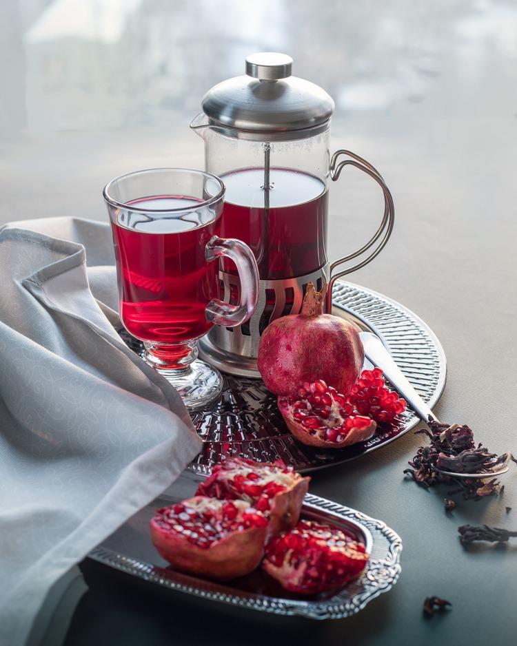 A imagem mostra uma jarra e um copo contendo chá de romã ao lado de dois frutos cortados pela metade.