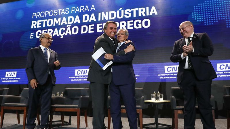 O presidente Jair Bolsonaro no palco de um evento promovido pela Confederação Nacional da Indústria (CNI). Ele está abraçando o ministro da Economia, Paulo Guedes, e sendo aplaudido por industriais.